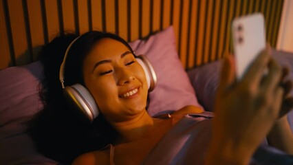 Happy Korean woman wearing headset lies in bed and texts on phone, social media