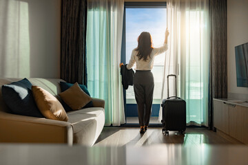 Confident businesswoman wearing white shirt and holding suit with suitcase stands in a hotel room while looking at the view from window Beautiful sunset. Woman enjoying her holiday in a luxury hotel.