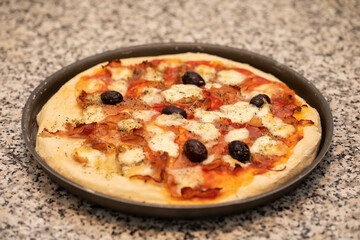 pizza fatta in casa, condita con pomodoro, olive nere, mozzarella e prosciutto crudo, in una teglia scura di metallo, su una superficie di marmo grigio