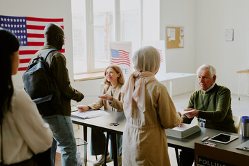 Mature Caucasian man and woman working at voting station giving ballot papers to multi-ethnic citizens, copy space
