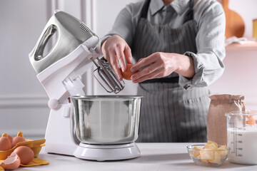 Woman adding egg into bowl of stand mixer while making dough at table indoors, closeup