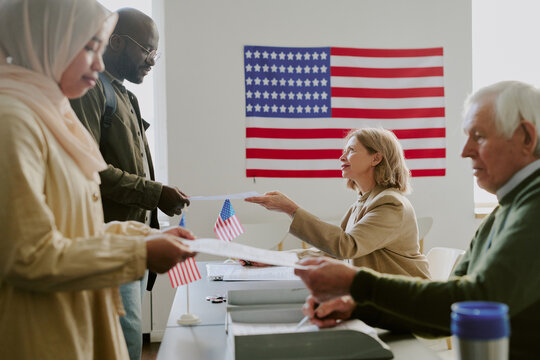 Side view shot of senior polling station workers giving ballot papers to ethnically diverse young voters