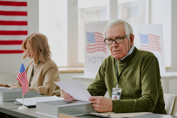 Senior Caucasian man holding ballot paper sitting at desk next to his female colleague at polling...