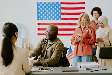 Cheerful young African American man in wheelchair taking ballot paper at polling place on election day, copy space