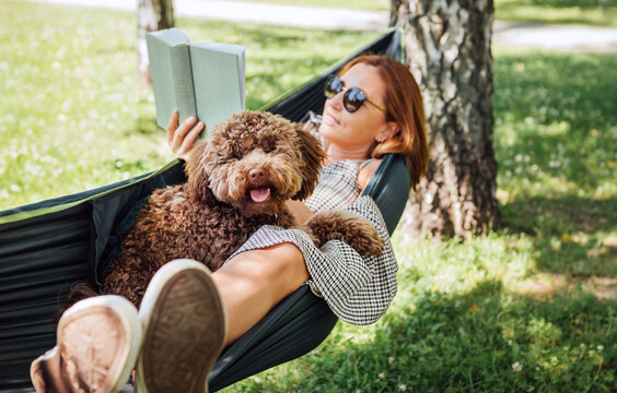 Woman reading book relaxing in hammock with her fluffy brown Maltipoo dog on sunny day. Both looking content and happy. This outdoor scene captures joy of bonding with pets and enjoying togetherness