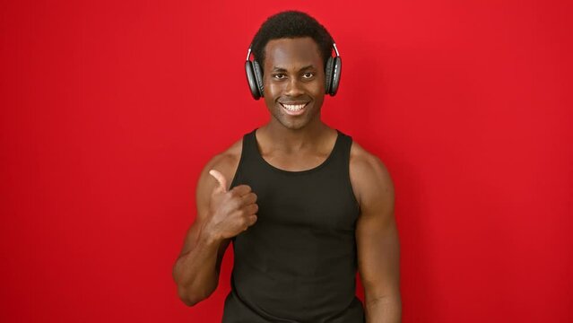 Joyful, confident young african american man listening to music, giving an enthusiastic thumbs up, pointing offside with a big open-mouthed smile, over a vibrant isolated red background.
