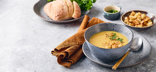 Bowl of mashed potato soup with a spoon on the plate. The bowl is filled with yellow soup puree and topped with croutons, parsley and ground red pepper.