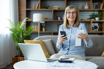 Smiling senior woman managing personal finances at home, using a smartphone while holding a...