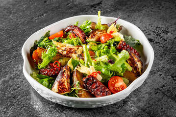Fresh mixed salad with grilled octopus, cherry tomatoes, and greens in a white bowl on a dark textured background