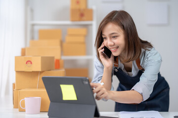 Young woman in a home office environment managing online orders on a tablet while talking on the phone, with packaging boxes in the background.