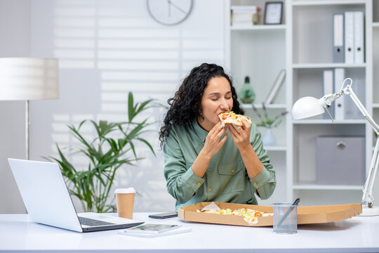 A woman eating pizza at her desk during a work break in a modern office setting. The scene captures a relaxed moment with a casual meal.