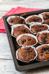 Tray of freshly baked chocolate muffins with chocolate chips on top, placed on a red cloth and white wooden table.