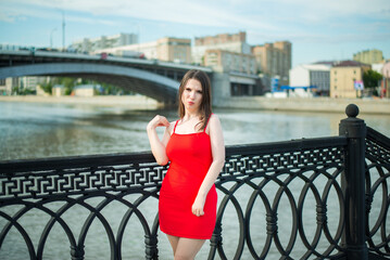 A woman in a red short dress stands on the embankment against the backdrop of a bridge over the river.