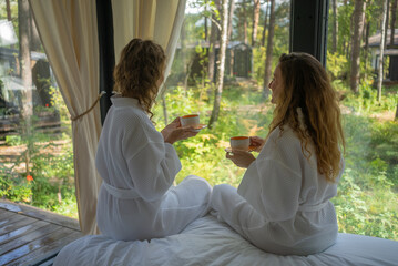 Two women in white robes sipping tea while sitting by a window with forest view, enjoying a...