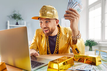 Rich man in a golden suit holding a stack of cash money and using a laptop, celebrating financial...
