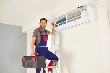 Portrait of professional electrician man or technician with screwdriver standing on ladder,looking at camera and smiling maintaining, cleaning, repairing or installing modern air conditioner indoors.