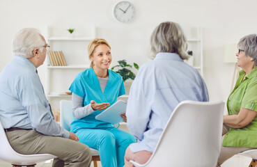Obraz premium Senior people at retirement home having meeting with nurse or doctor. Cheerful friendly young girl in scrubs with clipboard talking to group of old men and women. Senior care concept