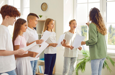 Portrait of a young school children standing in a circle with their young female teacher having music lesson in the classroom. Kids singing in a choir holding white sheets preparing for performance.