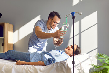Friendly male nurse gives IV drip to happy female patient undergoing vitamin therapy. Smiling man in light blue scrubs sets intravenous line for cheerful young woman lying on bed in medical office 