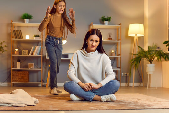 Frustrated, disappointed, tired, desperate child crying and having argument with mother. Mom sitting on floor at home, listening to disobedient teenage daughter and pretending to ignore her angry rant
