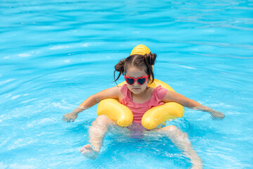 A little girl in sunglasses swims in an outdoor pool hugging an inflatable duckling. Sea holidays on vacation. Pretty female child smiles and swims along with inflatable yellow duck-shaped circle.