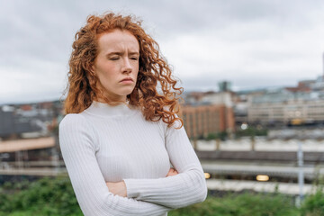 Unhappy Young Woman With Red Hair Stands With Arms Crossed in Front of Cityscape