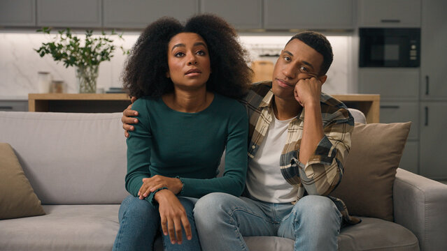 Annoyed African American ethnic exhausted young couple disturbed parents sitting sofa thinking feeling tired about noisy little boy hyperactive child misbehaving kid son playing run inside living room
