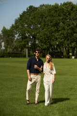 Fototapeta premium A beautiful young couple in elegant attire stands together in a lush green field, embodying an old-world luxury lifestyle.