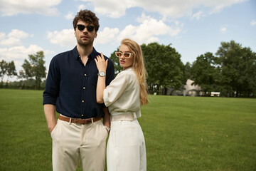 A chic young couple in elegant attire striking poses among lush greenery in a field.