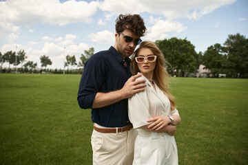 Fototapeta premium A man and woman, elegantly dressed, stand together in a lush green field, embodying a life of luxury and sophistication.