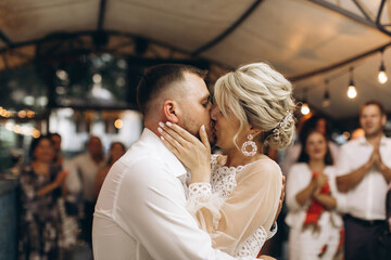 Bride and groom dancing the first wedding dance and kisses against the background of guests in an evening restaurant