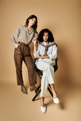 Two young, beautiful multicultural women in stylish attire, sitting on a chair, striking a pose for a picture.