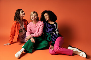 Three young women in vibrant attire showcase friendship and cultural diversity on an orange backdrop.