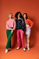 Fototapeta premium Three young beautiful multicultural women in stylish vibrant clothes stand united on an orange background.