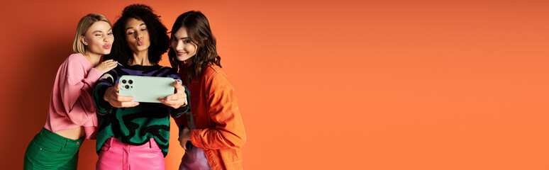 Group of diverse young women in stylish clothes posing for selfie together on orange background.