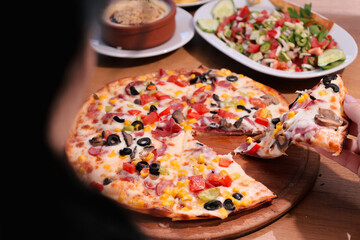 A hand holding a slice of vegetable pizza over a wooden table, featuring an array of toppings like olives and peppers.