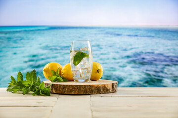 Lemon fruits, fresh mineral water, ice in glass on wooden table top with beautiful blur landscape of calm blue sea and blue sky. Vacation time and cool drink for hot day.