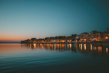 Obraz premium A serene view of a coastal town at dusk, with the city lights reflecting in the calm waters