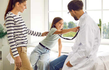 Obraz premium Family doctor lets kid patient have fun in his office. Mother and child visit doctor together. Happy little girl puts stethoscope to chest of her pediatrician. Health care, medical check up concept