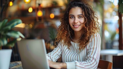 A young woman with curly hair is smiling while working on her laptop at a modern cafe, surrounded by plants and warm ambient lighting, giving an artistic appeal to the scene.