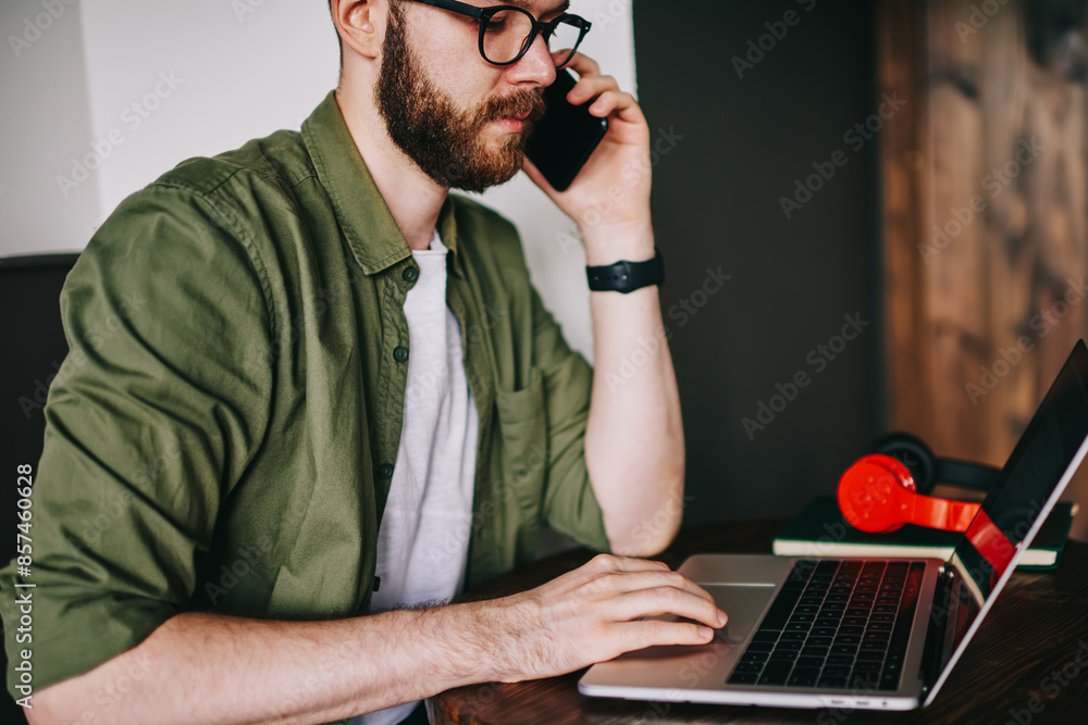 Poster stylish bearded hipster male talking by smartphone with girlfriend and using laptop in coworking spa
