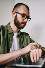 Young handsome man checking time on his smartwatch while chatting via laptop at favourite cafe,...