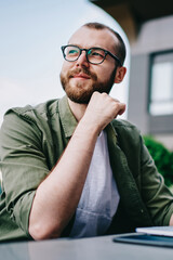 Young handsome male 20 years old in eyeglasses sitting at street cafe during free time with new modern laptop, bearded man freelancer  thoughtfully looking aside and thinking about distance work