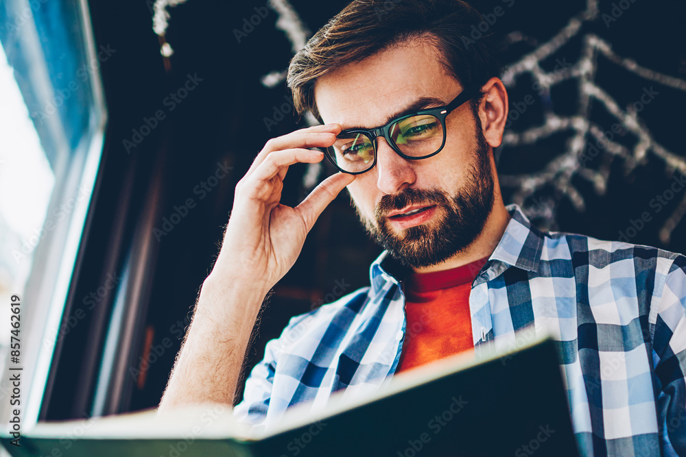 Poster concentrated smart young man with beard and optical eyeglasses searching useful information in textb