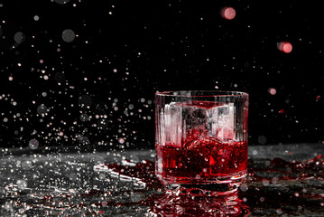 A close-up of a glass with red liquid and ice cubes, splashes around, against a dark background with light bokeh.