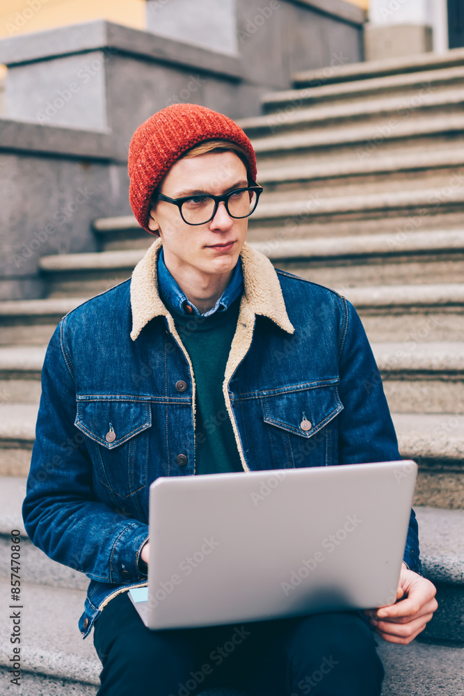 Poster thoughtful hipster guy blogger looking away while updating software on laptop computer via high spee