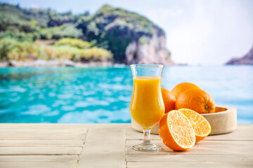 Orange fruit and fresh orange juice in glass on wooden table top with beautiful blur landscape of calm blue sea and blue sky and rocky mountain. Vacation time and cool drink for hot 