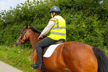 Close up shot of female rider wearing hi viz safety gear, riding her bay horse along a country lane...