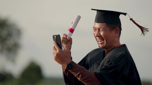 A man in a graduation gown is holding a cell phone and laughing. He is taking a picture of himself with his graduation cap and gown