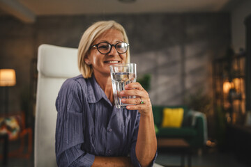 Portrait of mature woman sit, smile and hold glass of water at home